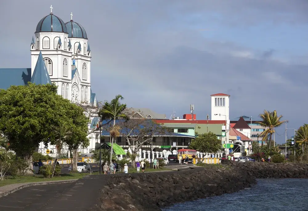 Sunny morning with slightly cloudy skies pictured from the walking path by the white cathedral built on a rocky black coast during the cheapest time to visit Samoa