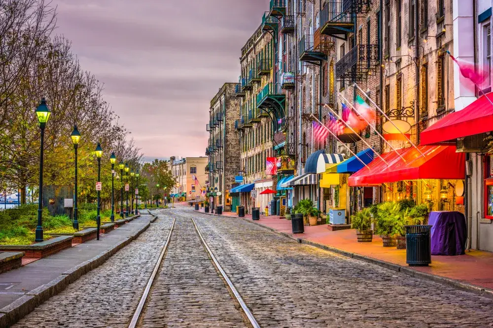 Savannah GA pictured at night with lights on the shops on the riverfront with the person walking along the tracks for a piece on whether or not Georgia is safe to visit