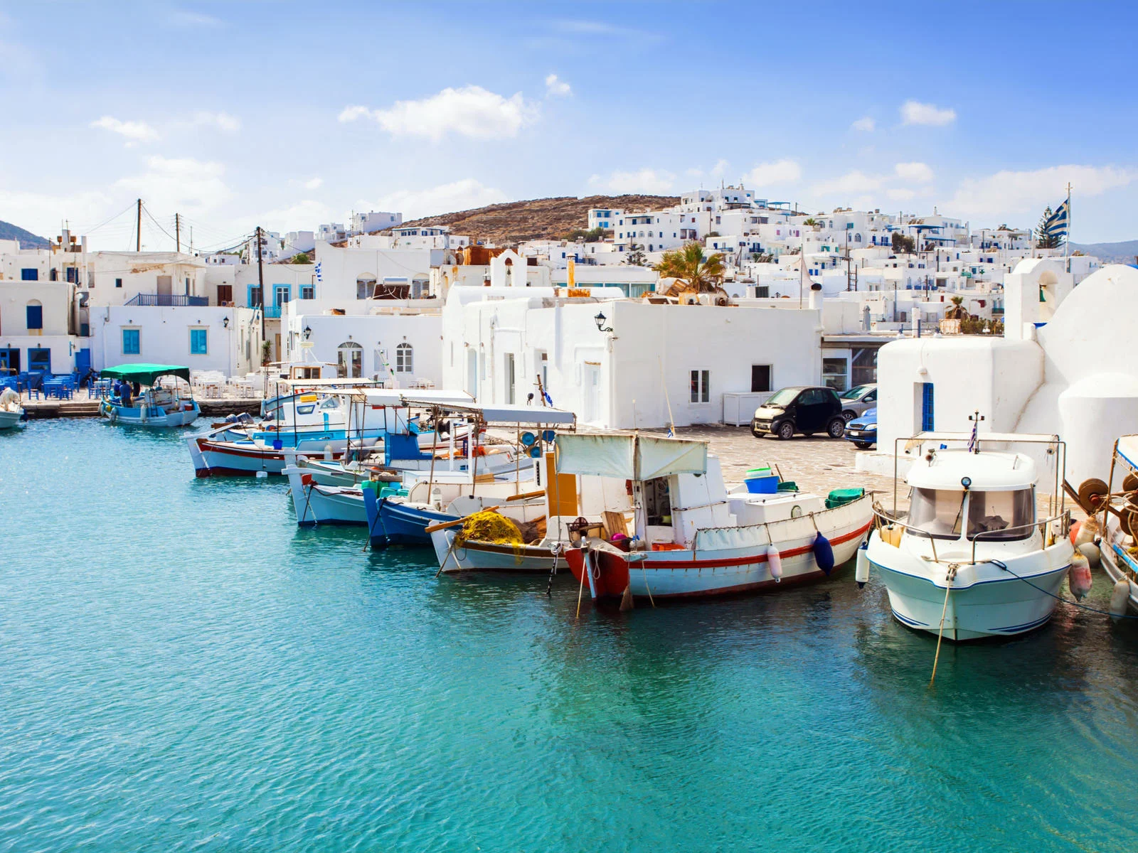 Several fishing boats docked at the coast of Naousa Village at Paros Island, one of the best islands in Greece to visit, where cars are parked near its signature white houses