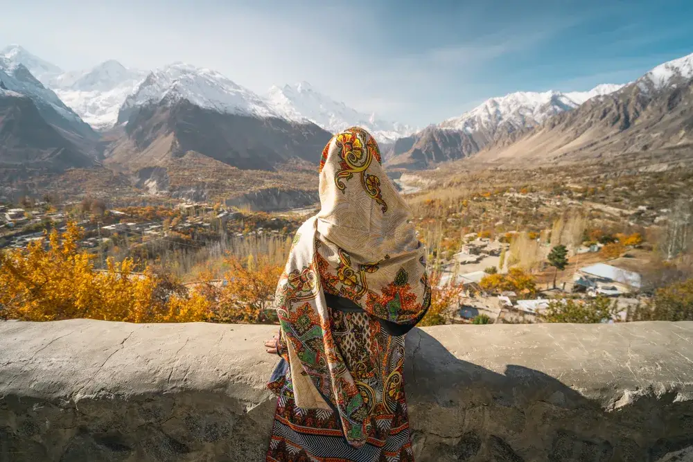 A woman wearing traditional dress sitting on wall and looking at Hunza valley in autumn season, Gilgit Baltistan in Pakistan, Asia