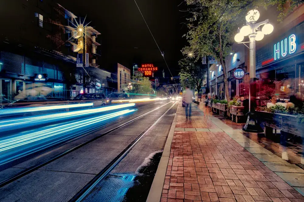 Very cool low-exposure image with cars driving by a bustling paver sidewalk with lots of little shops and restaurants lining the walkway, as seen at night during the best time to visit Tucson