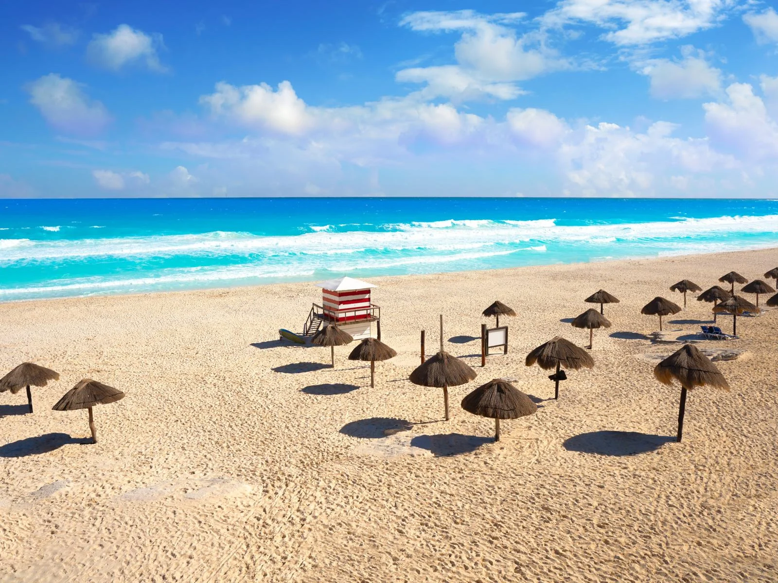 Single pole tiki huts lined up at the vast shore of Playa Delfines, a pick on the best beaches in Cancun, and foamy blue sea due to crashing waves