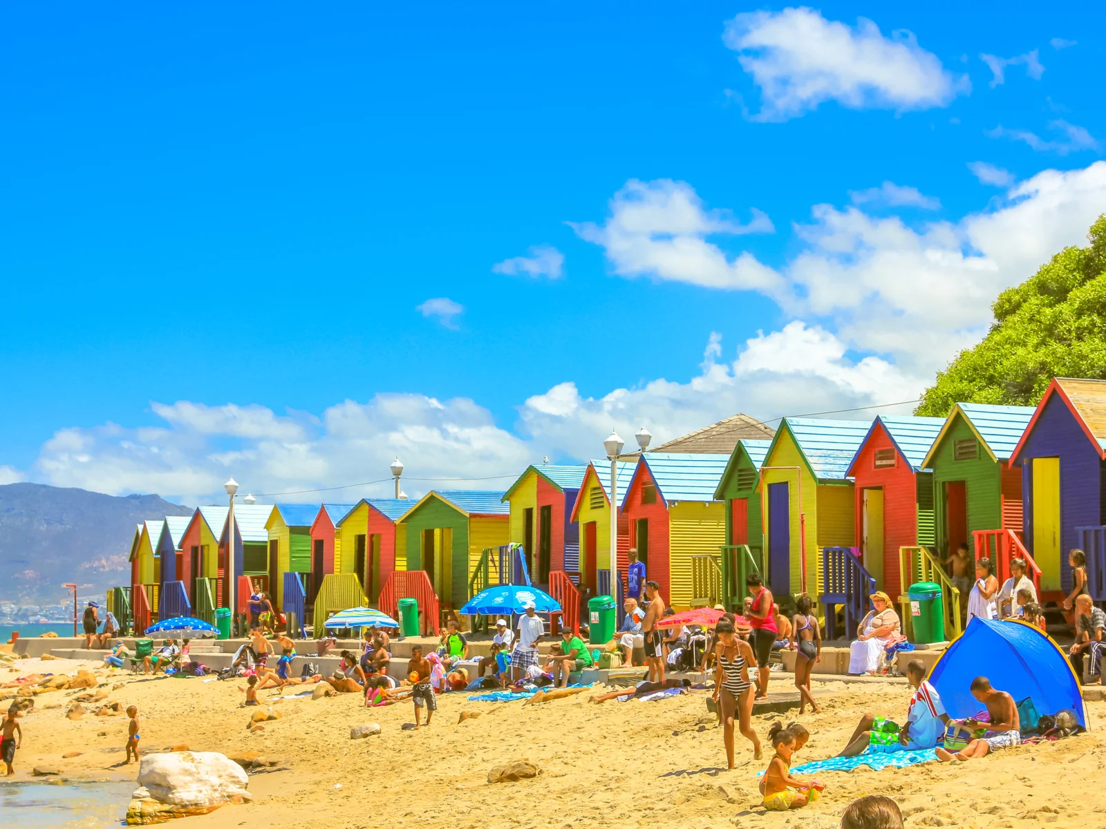 Gorgeous sunny day view of a beach in Cape Town pictured in front of multicolored houses for a piece titled Is South Africa Safe to Visit