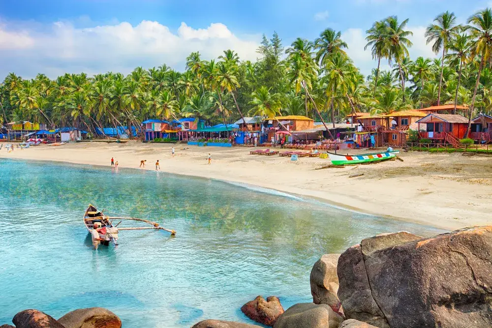 Boat on the water with a man wading next to it in front of small cottages on the beach during the best time to visit India