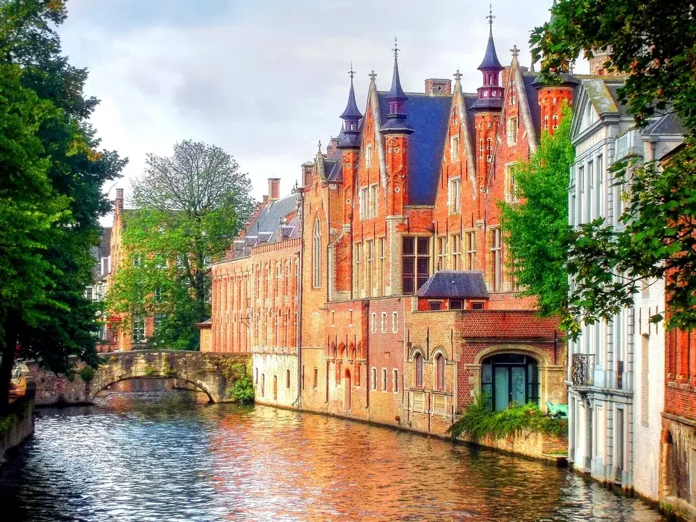 Bruges canal pictured from a walking path looking down toward a bridge with a cloudy sky and old brick buildings on the right during the overall best time to to go Belgium