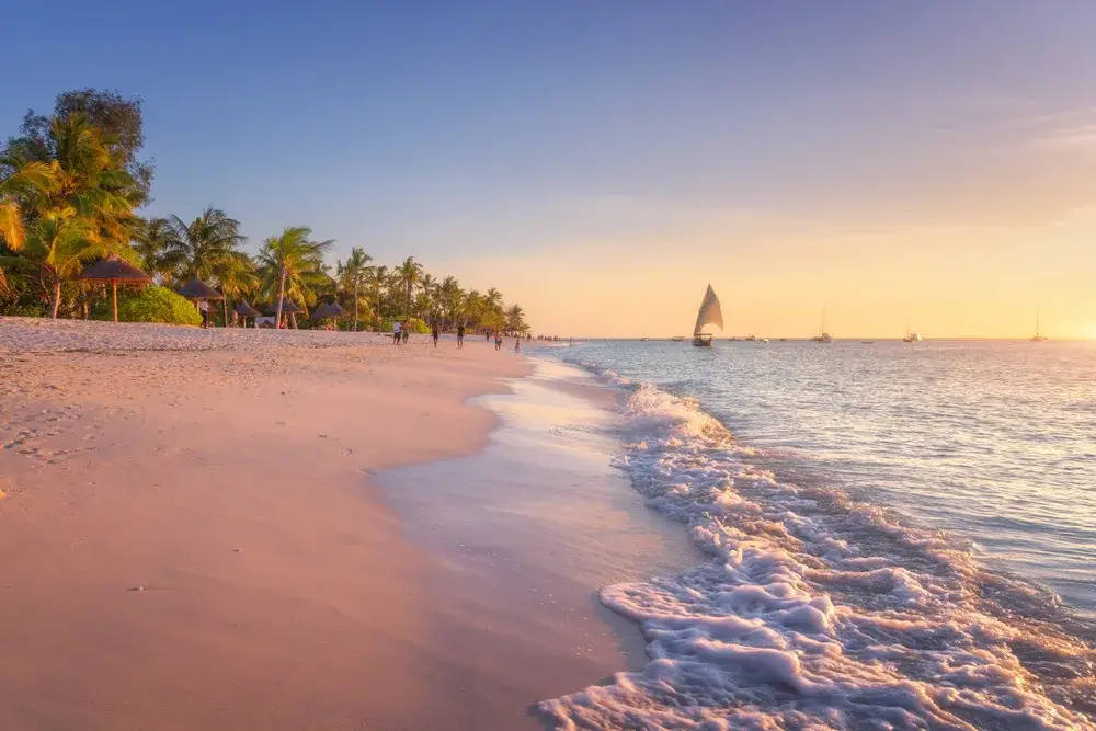 Long expansive stretch of beach pictured at dusk with a sailboat in the background taken during the cheapest time to visit Zanzibar